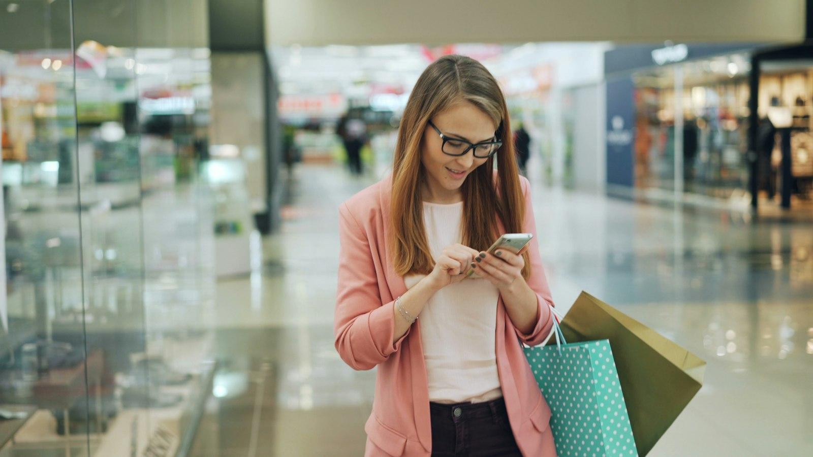 A woman checking her phone in a shopping mall while carrying shopping bags