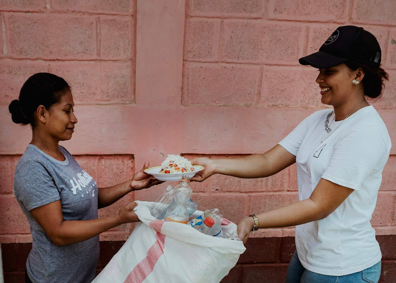 A woman hands a plate of food to another woman outside a pink wall