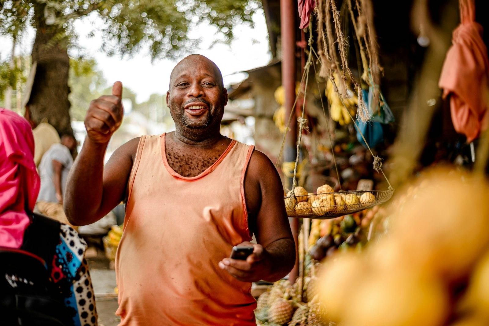 A smiling man holding his phone in front of a fruit stall at an outdoor market